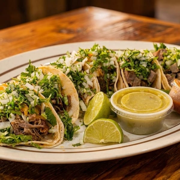 A white oval plate with five street tacos, lime wedges, and two small sauce containers on a wooden table.