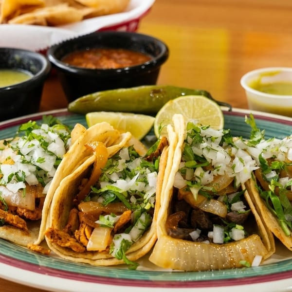 A plate of four street tacos with various fillings, cilantro, and onions, with salsas and lime.