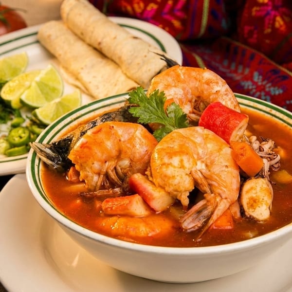 A bowl of shrimp and seafood soup with tortillas, salsa, and limes on the side.